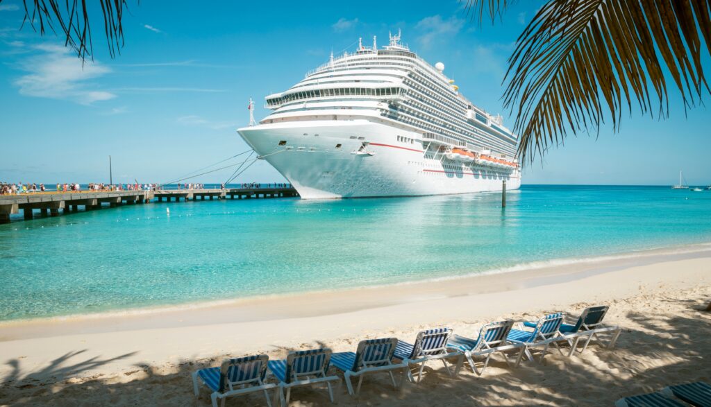 Cruise ship docked near tropical beach with clear blue water and palm trees.