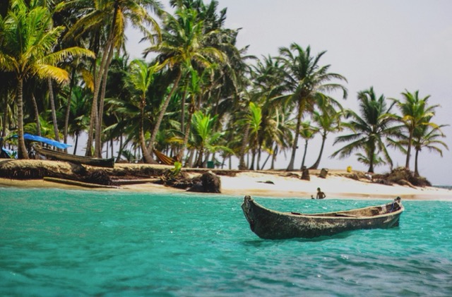 Small wooden boat floating on clear turquoise water near a tropical palm-lined island.